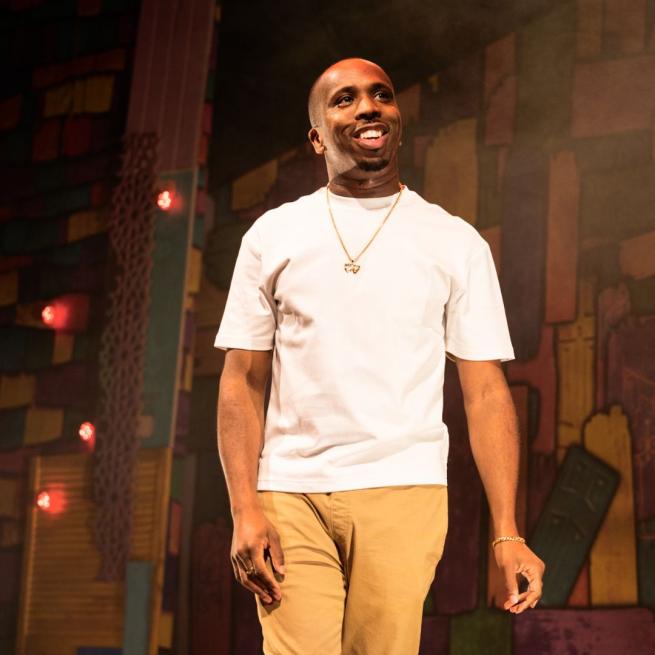 Kiell Smith Bynoe smiles onstage, wearing a white t-shirt and gold pendant chain under warm lighting.