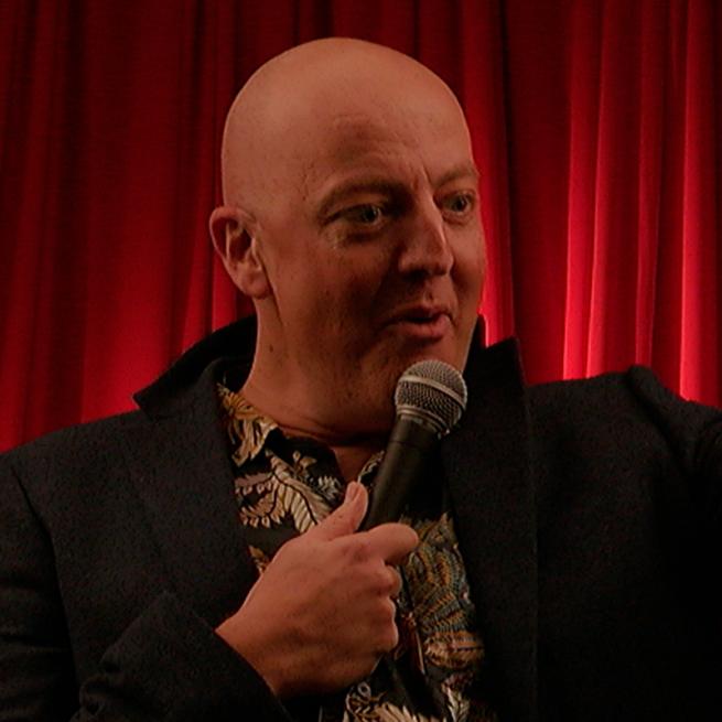 Antony Vila holding a microphone on stage in front of a red curtain.