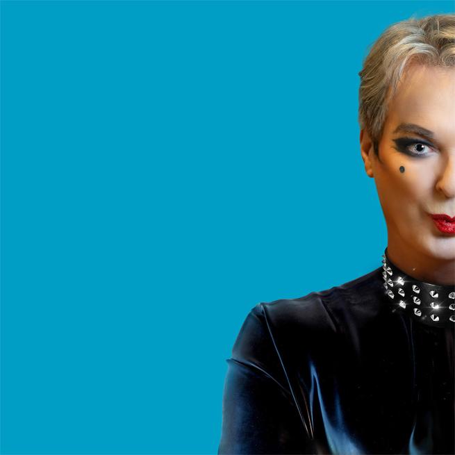 Half of Julian Clary's face is visible, pouting with red lipstick, smoky winged eyeliner and a beauty spot. He also wears a studded leather collar.