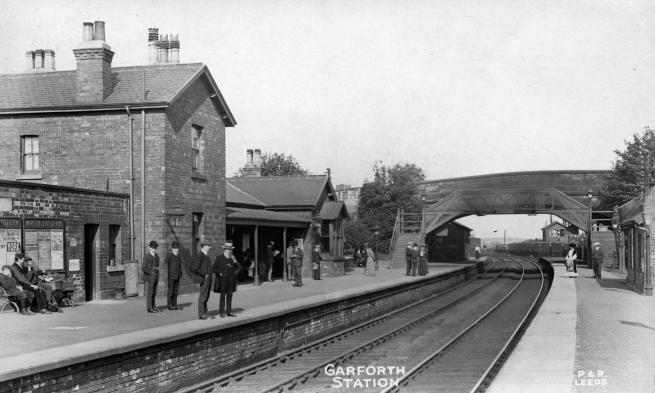 Historic black-and-white photograph of Garforth railway station. Brick station buildings line the left platform, where several people stand and sit beneath railway posters. Curving railway tracks run through the centre of the scene beneath a covered footbridge linking the platforms. Text at the bottom of the photograph reads “GARFORTH STATION”.