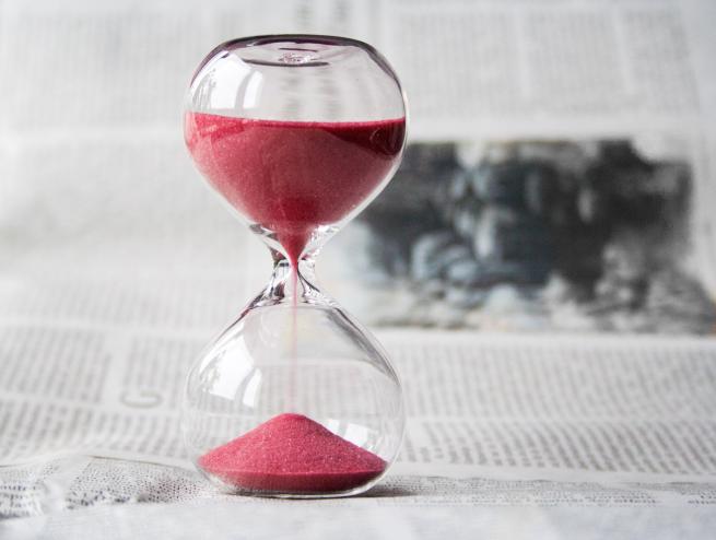 Close-up of a modern glass hourglass with pink sand against a blurred background of newspaper text, symbolizing the passage of time or a deadline.