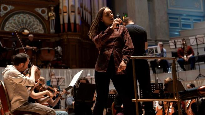 A singer holding a microphone performs onstage in front of a live orchestra and pipe organ inside a grand concert hall.