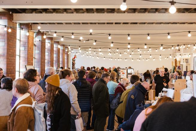 A crowd of visitors exploring the Christmas Market in the 1912 Mill event space. The room is a former industrial textile mill, with red brick walls and strings of bright festoon lights hanging across white steel beams. 