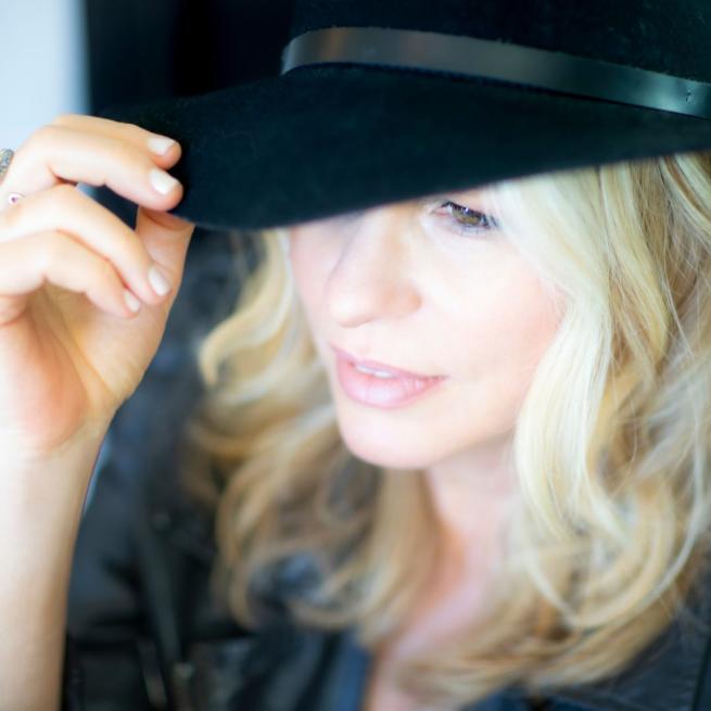 Vonda Shepard lightly tips the brim of her black hat, from a candid camera angle.