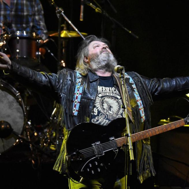 A bearded musician stands on stage with a microphone and electric guitar, arms outstretched.