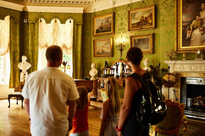 Four figures looking into a historic room, with a piano, fireplace and painted portraits.