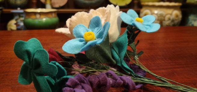 A picture of blue white and green felt flowers on a wooden counter top with art pottery in the background.