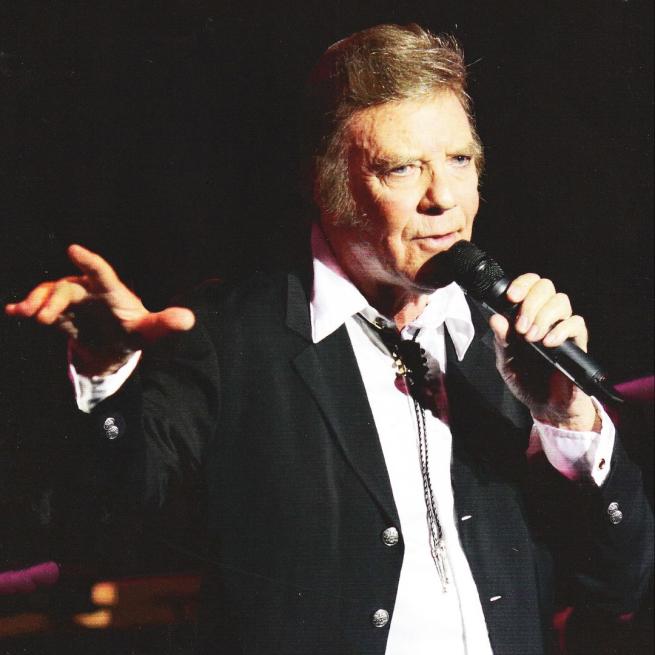 Marty Wilde sings into a microphone on stage, wearing a suit and bolo tie.