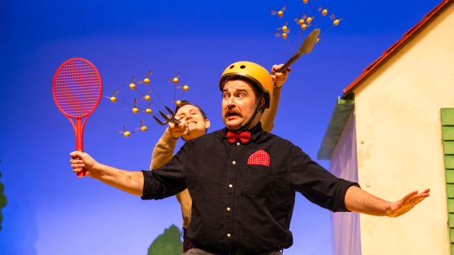Two performers acting on stage. One is holding a tennis racket and the other is holding branches with leaves on them. 