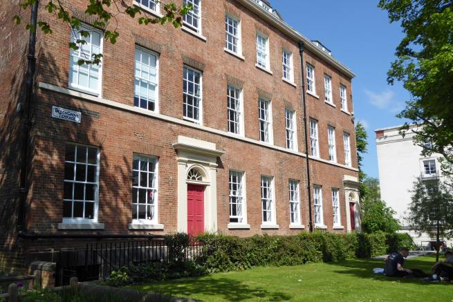 Three-storey red-brick Georgian terrace with white sash windows and red entrance doors, viewed from a grassed garden. Trees frame the building, and a street sign on the corner reads “Beech Grove Terrace”. Two people sit on the lawn at the right-hand side of the scene.
