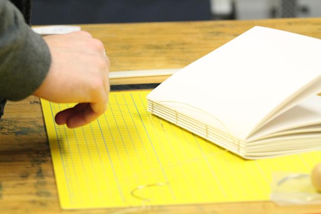 A hand reaching out to a coptic bound white notebook, which is positioned on a bright yellow cutting mat on a wooden table.