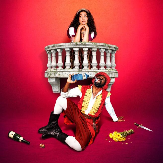 Stood at a prop balcony, an actress in period costume rests her head in her hands and looks off-camera. In front of her, another actor is sat on the floor in a red tunic and boots, drinking from a bottle of gin beside an empty bottle and discarded dagger.