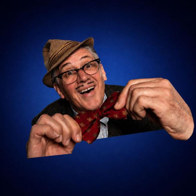 With comically large hands holding onto a big bow-tie, Count Arthur Strong smiles, wearing a brown trilby hat and glasses.