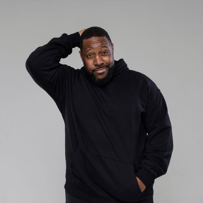 Aurie Styla smirks, with one hand held behind his head, the other in the pocket of his black hoodie, pictured against a grey backdrop.