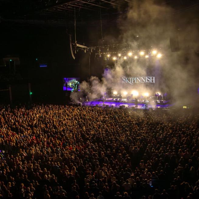 A packed crowd of people watch Skipinnish perform on stage, accompanied by spotlights and haze.