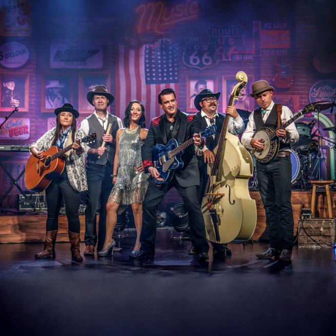 The members of A Country Night in Nashville stand together on stage in period costume, each holding their guitar or instrument. American memorabilia, photographs and flags are visible in the background.