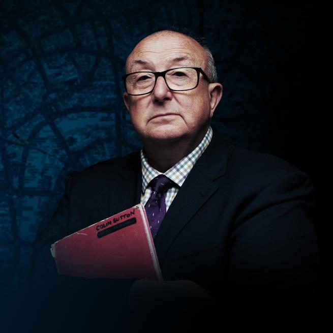 Colin Sutton in a suit and glasses with arms folded around a red notebook.