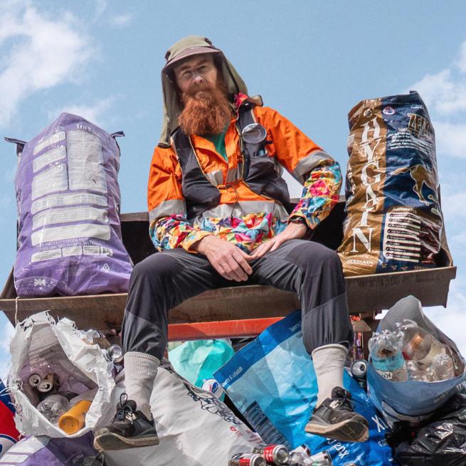A man with a long ginger beard, a floral shirt and orange high-vis jacket sits on top of a small mountain of bin bags containing cans and plastic bottles in front of a cloudy sky.