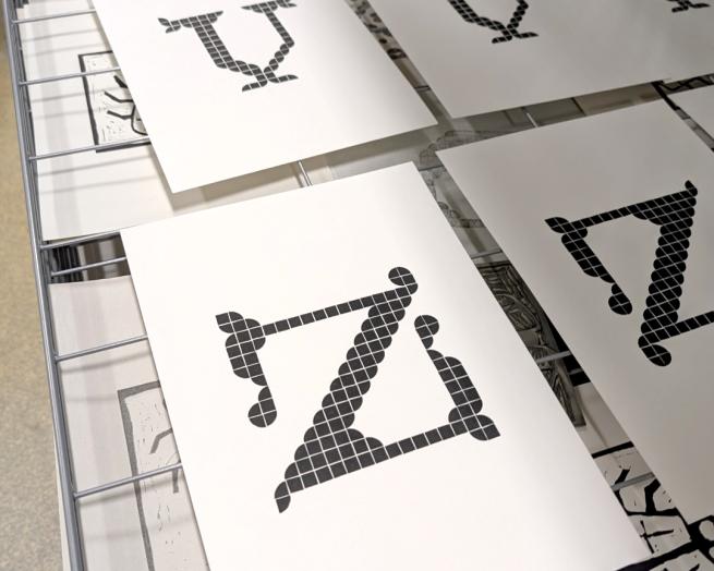Prints of a medieval style letters printed from lego plates. The prints are laid out in rows on a drying rack.