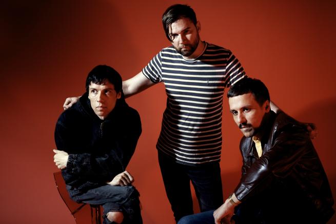The three members of the band The Cribs pose against a red background; one sits on a stool in a black jacket, another stands in a striped shirt, and the third sits in a brown leather jacket.
