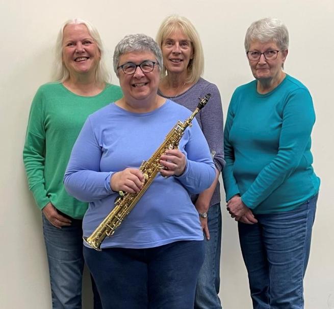 Four delightful looking ladies smiling for camera, one is holding a saxomophone