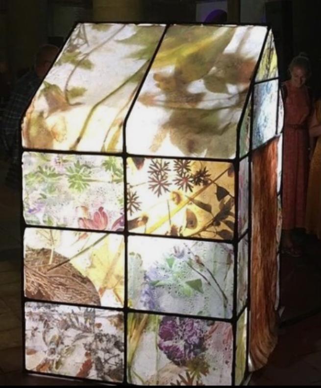 Image shows a piece of art in the shape of a greenhouse covered in images of flowers and lit up from inside.