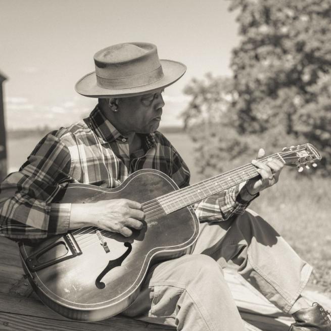 Black and white of Eric Bibb sat outdoors playing a guitar, wearing a plaid shirt and hat shading his face.