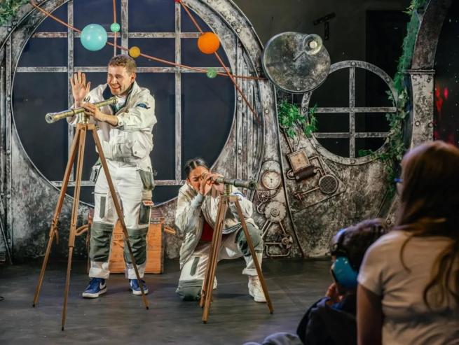 Two people looking through telescopes with a metallic space themed theatre set and hanging planets.