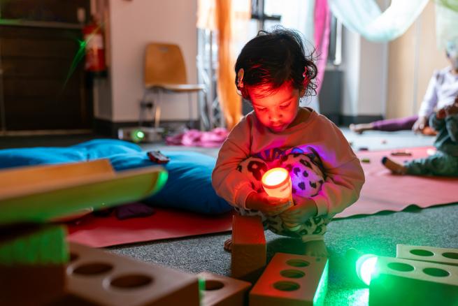 A young child crouches on the floor holding a glowing light, surrounded by illuminated sensory toys and soft furnishings.