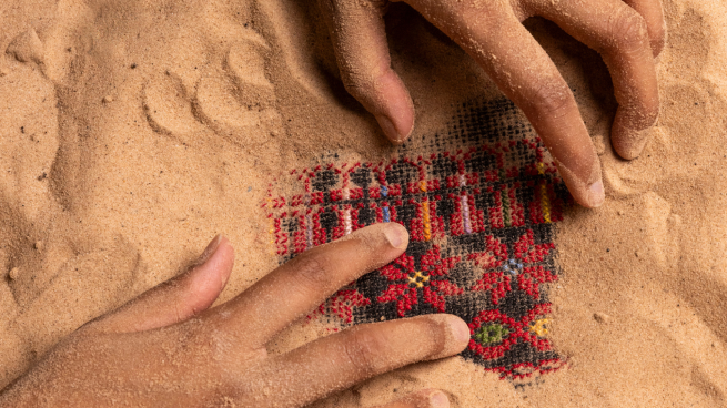 Two hands covered in sand are uncovering a piece of red fabric woven with flowers and a black and red repeating geometric pattern which is buried in the sand. 