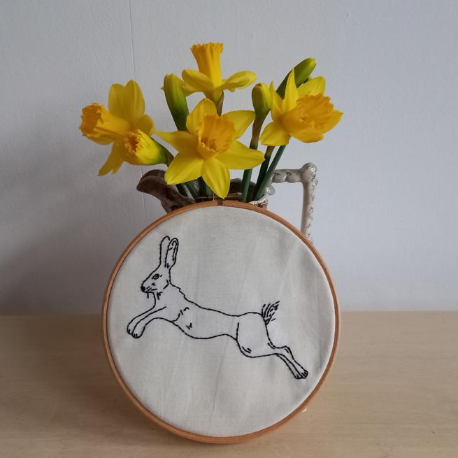 A photograph of an embroidery hoop with calico and a rabbit stitched in black thread. The hoop is popped up against a jug which holds yellow daffodils.