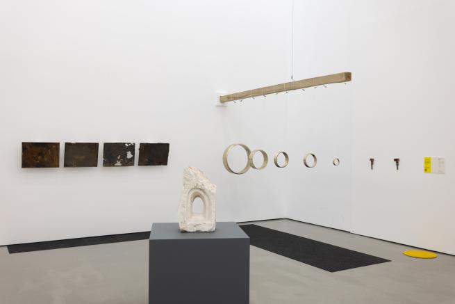 A gallery space with an abstract white marble sculpture in the foreground, a metal ring installation to the right, and rectangular bronze works attached to the wall in the background.