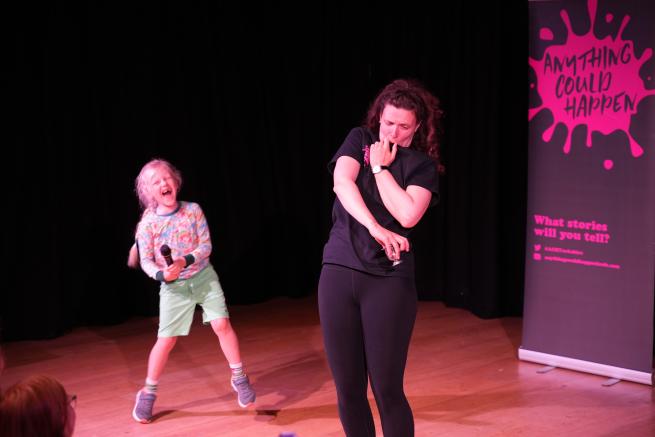 An 7 year old boy with a microphone laughs with delight as a woman strikes a funny pose on a stage