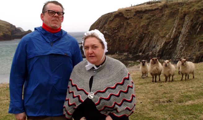 A man stands proudly in the bracing wind, he is adorned by a rather fetching blue cagool, the lady next to him has a white bonnet and knitted shawl. Behind is a harsh Nothern ladscape of sea cliffs and assorted sheep. Could be Wensleydale Sheep, though I'm no expert