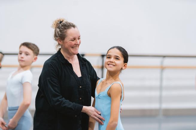 A dance teacher corrects a young dancer's posture. They are both smiling.