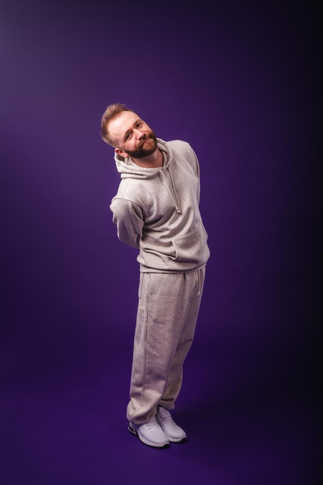 A white man with a beard wearing a full white tracksuit, leaning to the side and smiling in a warm but goofy manner on a purple background