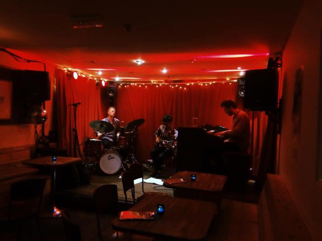 A guitarist, drummer and pianist play on a red lit stage against a red curtain.