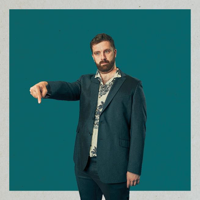 In a forest green suit and shirt patterned with palm trees, a straight-faced Pierre Novellie looks off-camera and points his finger downwards, set against a plain blue-green backdrop.