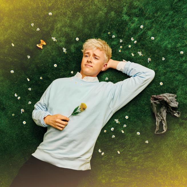 Daniel Foxx lies on a bed of soft grass and gazes at the sky, surrounded by daisies and holding a flower.