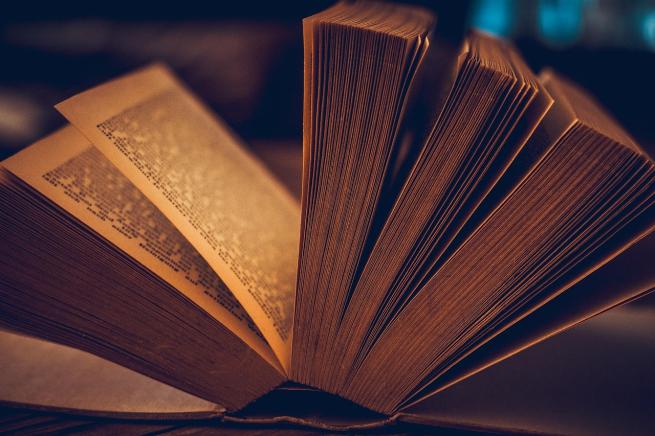 Close-up of an open book with its pages fanned out, warmly lit to highlight the texture and edges of the paper. The text on the visible page is small and densely packed but not readable due to the angle and focus.
