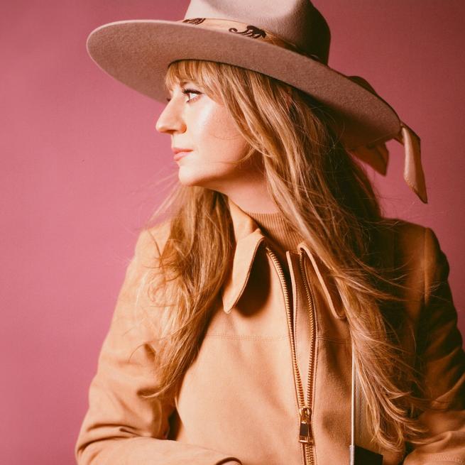 Joanne Shaw Taylor looks off to the side, holding a well-worn acoustic guitar and cowboy hat against a pink backdrop.