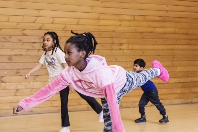 A girl is in a dance studio balancing on one leg, with the other extended behind her, her arms are stretched to the side
