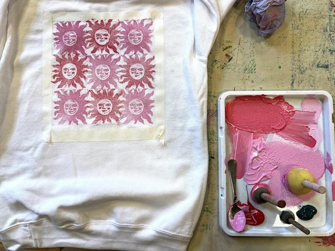 A white tshirt laid on a table with a repeat pattern of a sun freshly printed onto it. To the right is an ink palette filled with pink and red ink and sponge brushes