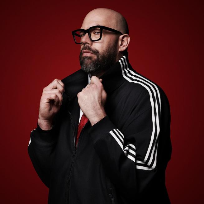 Tom Davis smoulders at the camera and tugs on the collar of his Adidas track jacket against a deep red background.