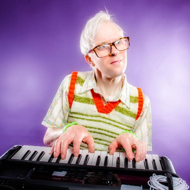 Robert White in glasses and a sweater vest over a patterned shirt, sat at a keyboard in front of a purple backdrop.