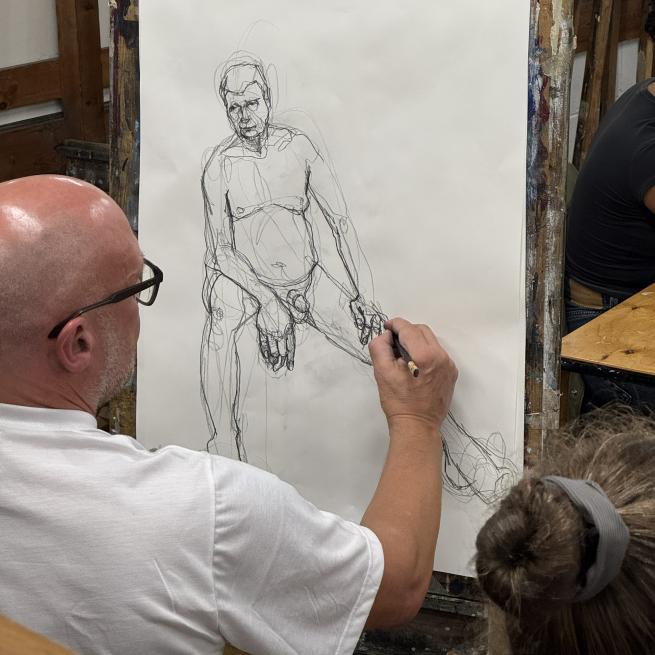 A photograph taken over someone's shoulder showing them drawing a male figure.