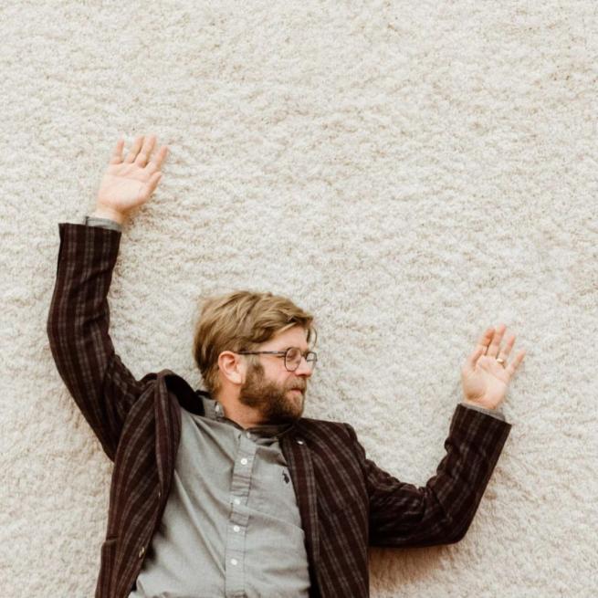 Chris Luedecke lying on a white rug, with arms outstretched and eyes closed. He wears glasses, a grey button-up shirt and chequered blazer.