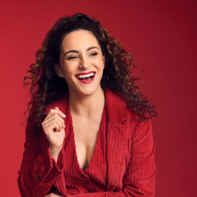 Janine Harouni in a red cord suit laughing against a red background.