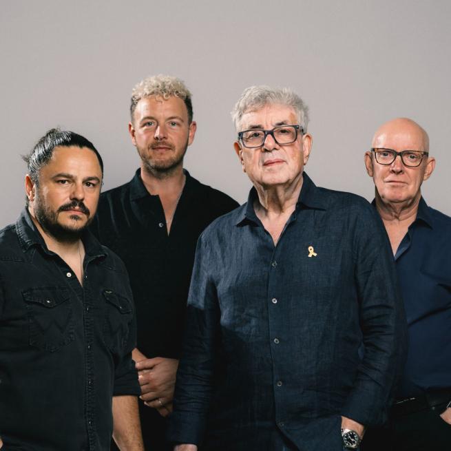 Graham Gouldman and Heart Full of Songs stand in front of a grey backdrop.
