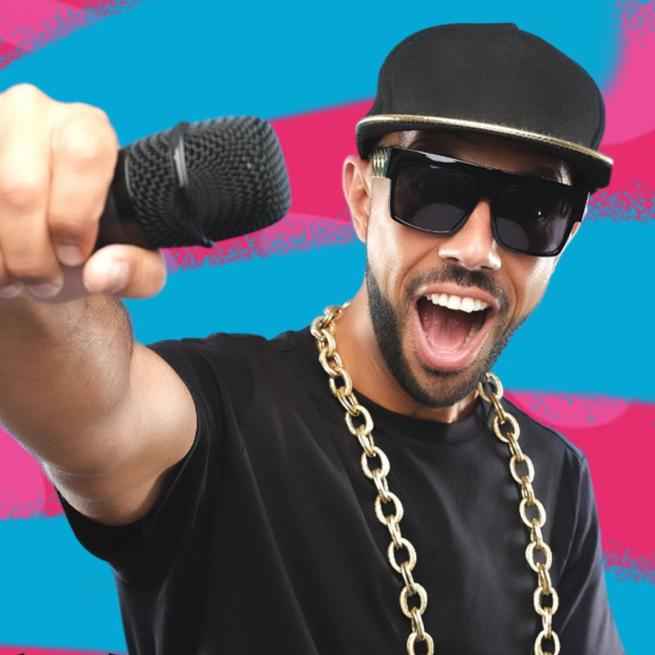 MC Grammar holds out a microphone in front of him, with a wide open-mouthed smile, wearing a black baseball cap, dark sunglasses and a thick chain, in front of a blue and pink graffiti background.